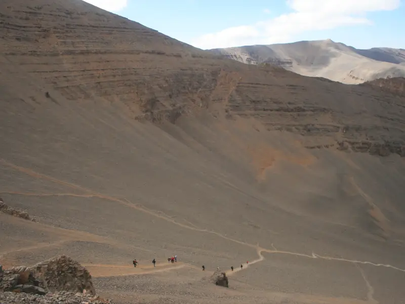 Eine Trekkinggruppe steigt auf einem schmalen Serpentinenpfad einen weiten Geröllhang im marokkanischen Mgoun-Massiv hinab.