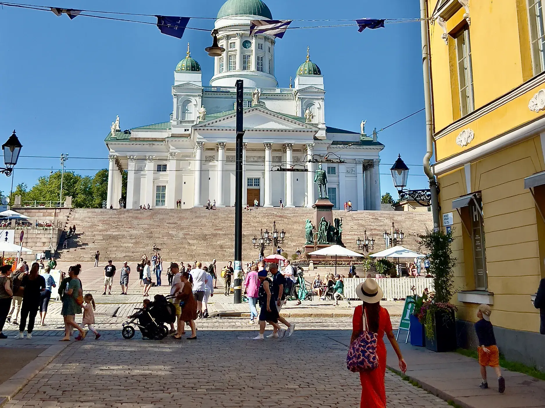 Zahlreiche Besucher flanieren bei Sonnenschein über den belebten Senatsplatz vor dem imposanten, weißen Dom von Helsinki.