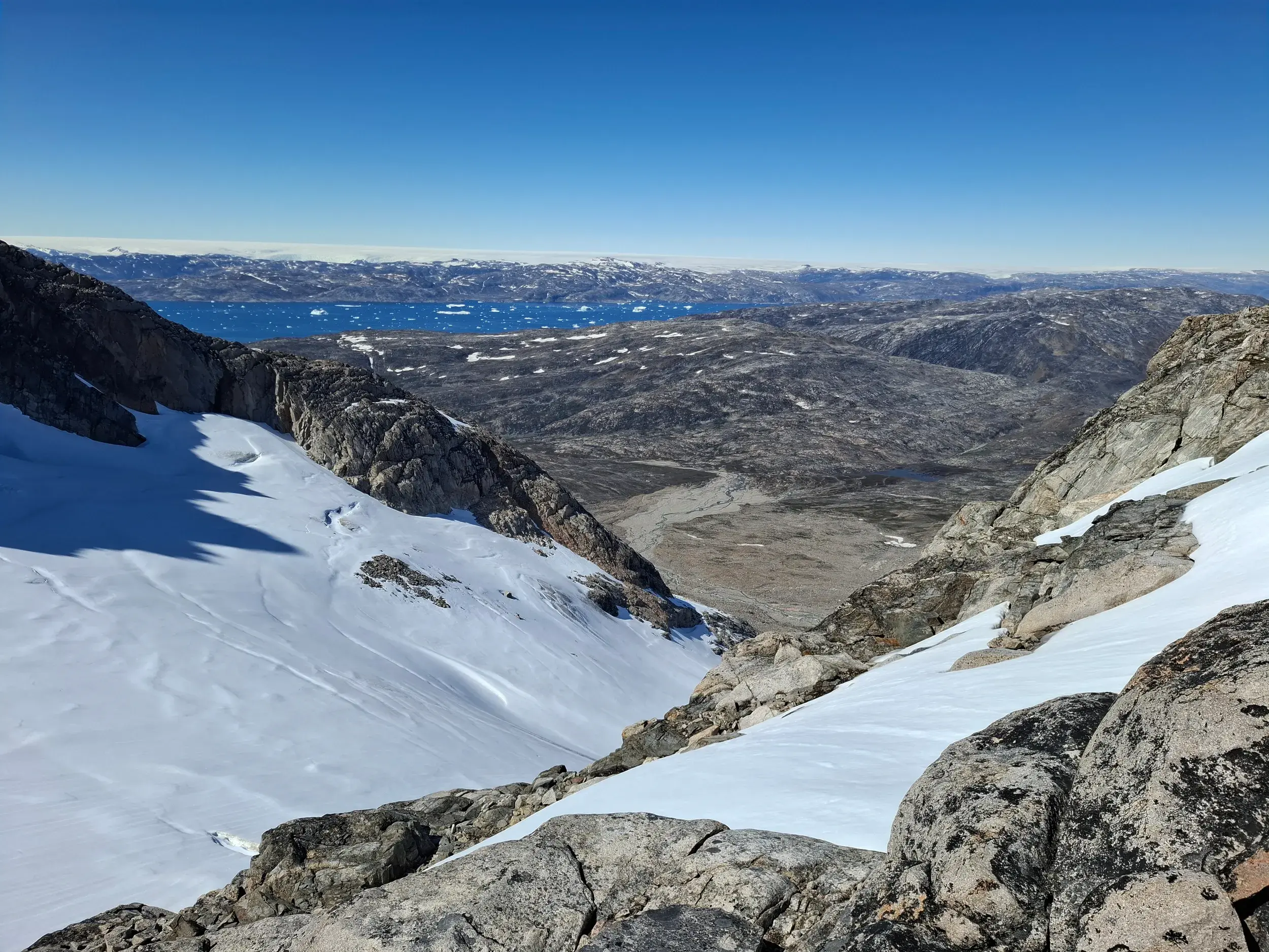 Weitblick beim Trekking in Ostgrönland von einem felsigen, schneebedeckten Hang auf den weiten Sermilik-Fjord mit Eisbergen.