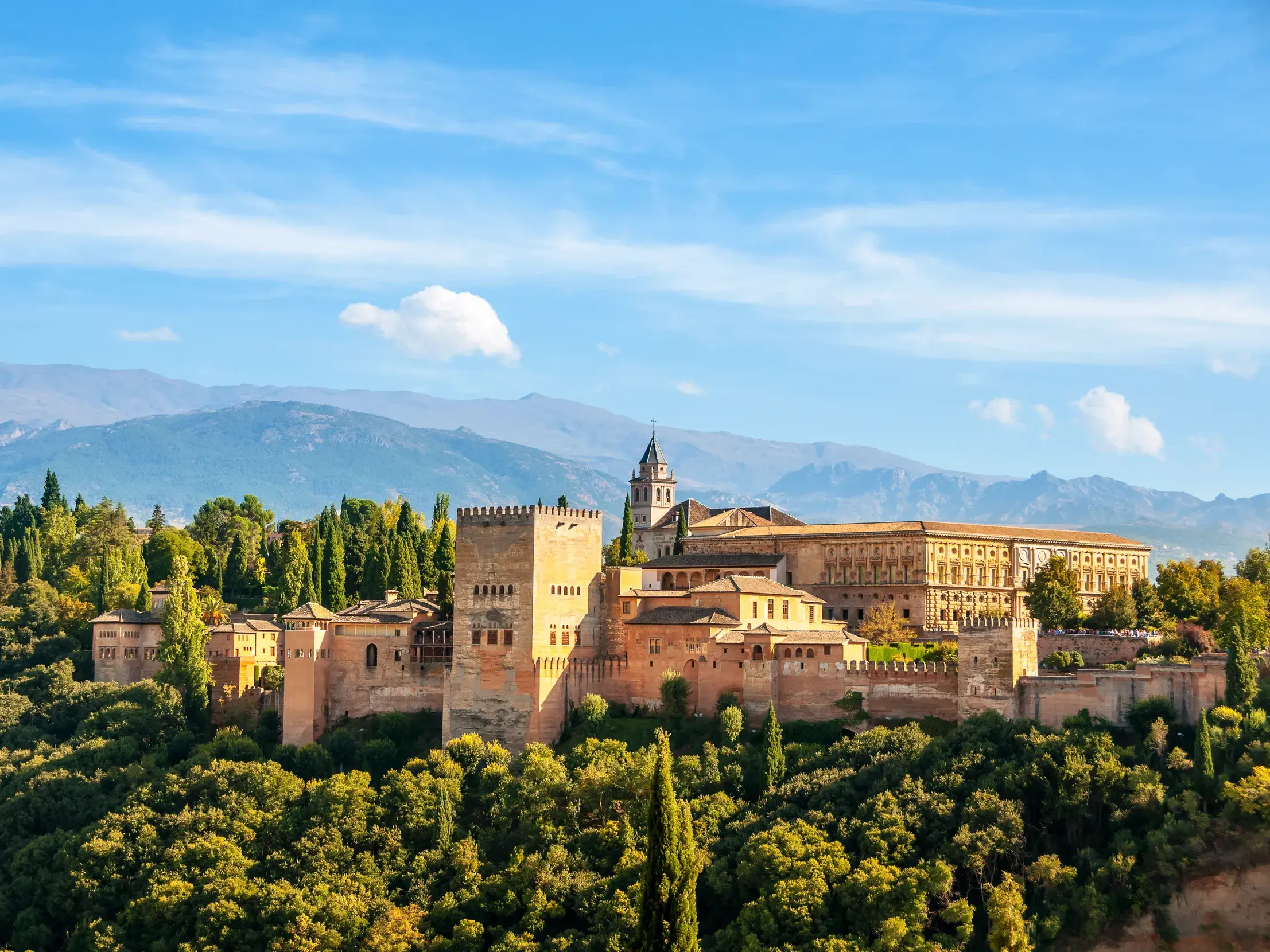 Die prächtige Burganlage der Alhambra in Granada auf einem bewaldeten Hügel vor der majestätischen Sierra Nevada unter blauem Himmel.