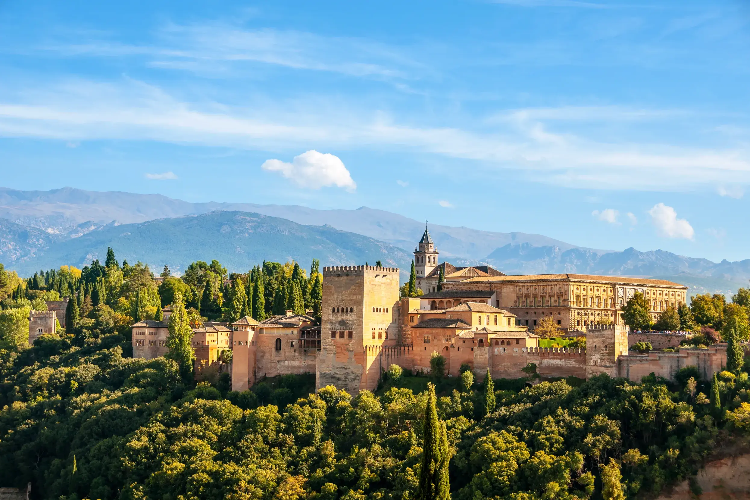 Die prächtige Burganlage der Alhambra in Granada auf einem bewaldeten Hügel vor der majestätischen Sierra Nevada unter blauem Himmel.