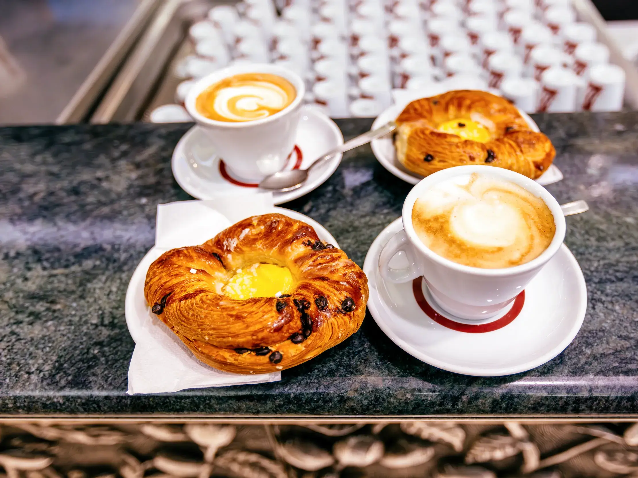 Zwei Tassen Cappuccino und süßes Gebäck für ein typisch italienisches Frühstück an einer Marmortheke in einem Café in Rom.