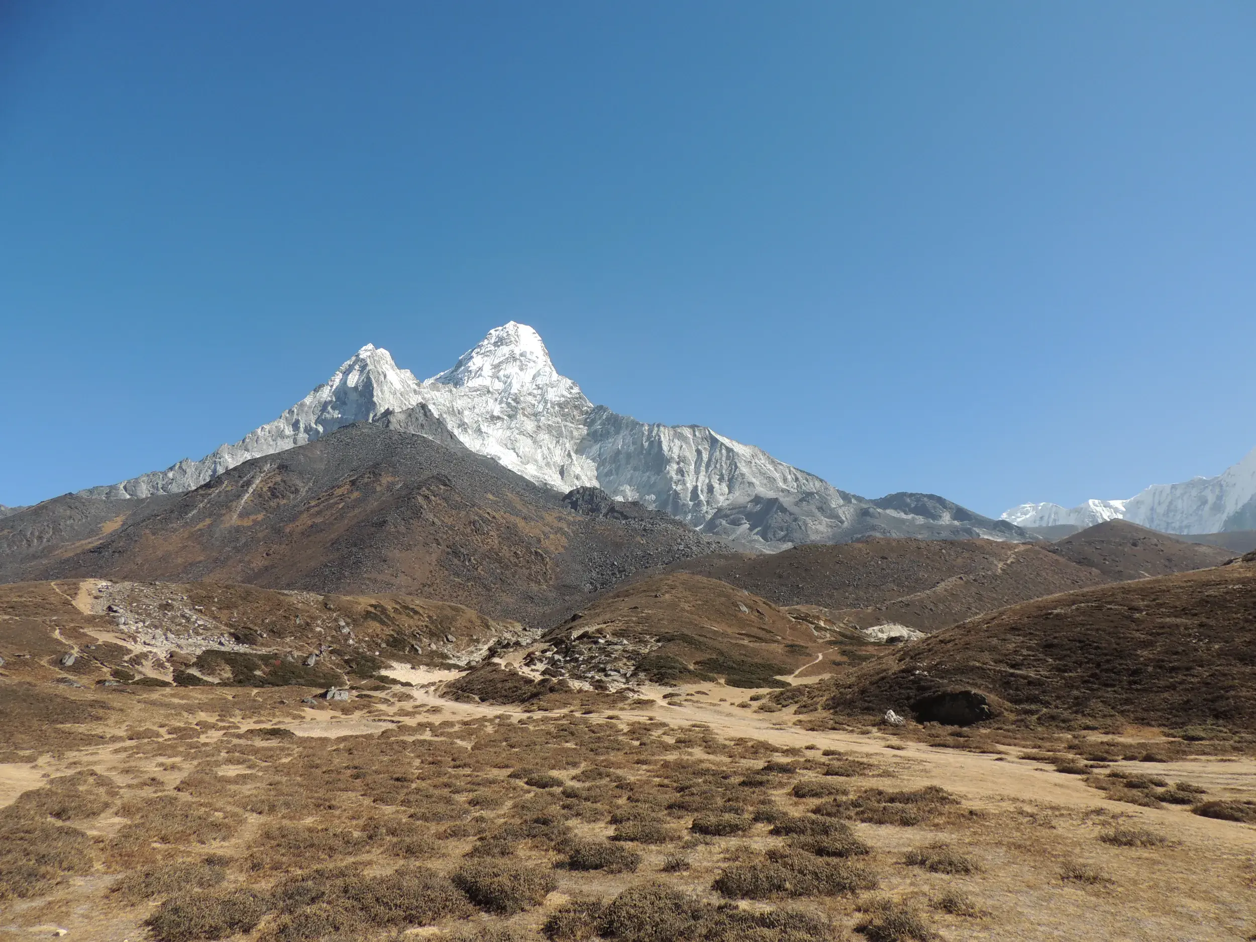 Karge Hügellandschaft mit Wanderpfad im Vordergrund, dahinter ragt der schneebedeckte Gipfel der Ama Dablam in den Himmel.