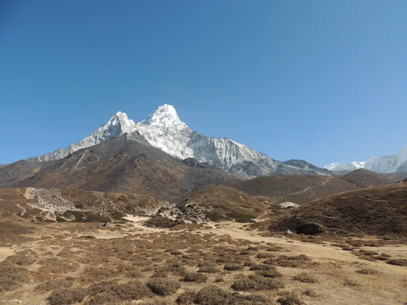 Karge Hügellandschaft mit Wanderpfad im Vordergrund, dahinter ragt der schneebedeckte Gipfel der Ama Dablam in den Himmel.
