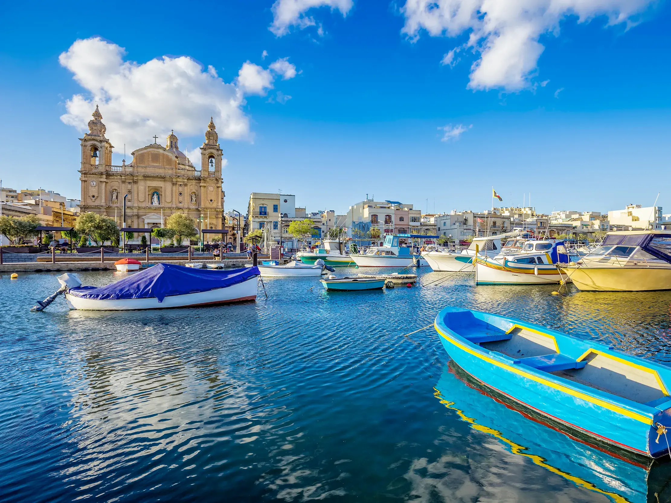 Bunte Fischerboote, auch Luzzi genannt, schaukeln im Hafenbecken von Marsaxlokk auf Malta vor der sonnigen Uferpromenade.