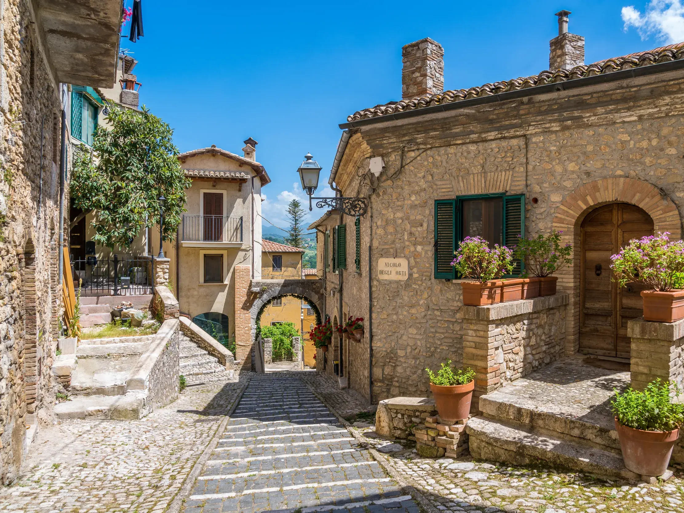Eine sonnige Gasse mit Kopfsteinpflaster führt durch das historische Bergdorf Labro in Latium, vorbei an alten Steinhäusern.