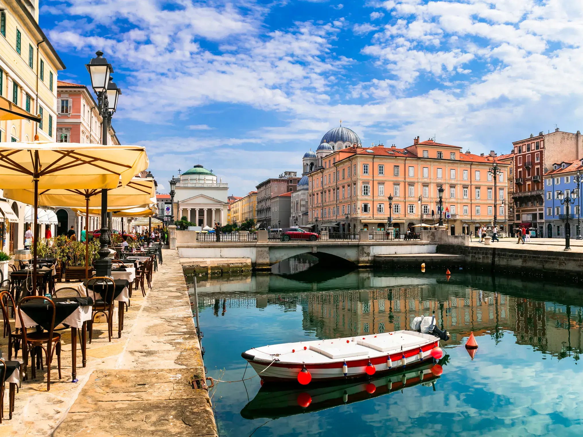 Ein Boot auf einem Kanal in Triest, gesäumt von Cafés und bunten, historischen Häusern unter einem sommerlichen Himmel.