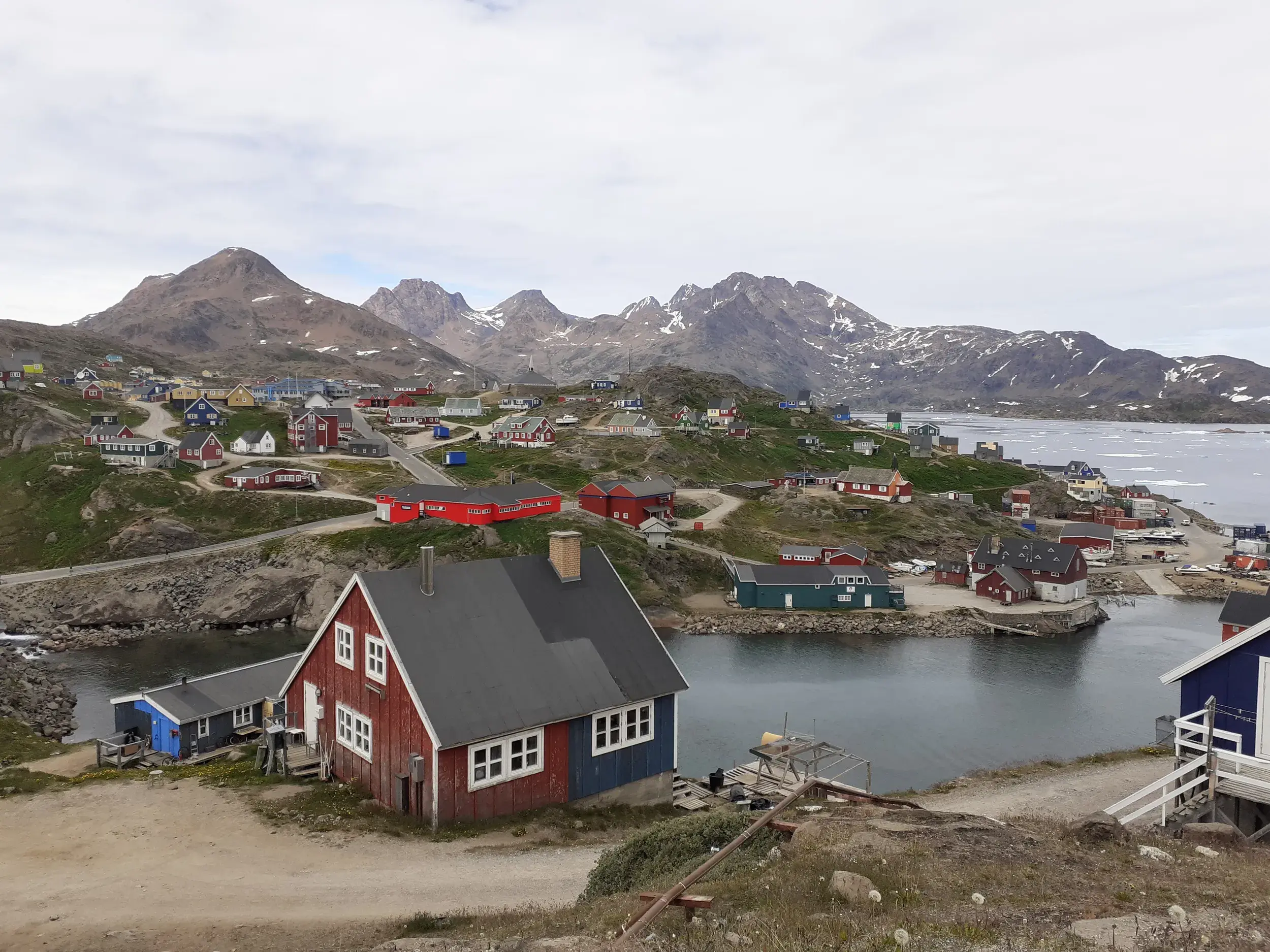 Bunte Holzhäuser der Siedlung Tasiilaq in Ostgrönland, gelegen an einer Bucht vor der rauen, felsigen Bergkulisse des Fjords.
