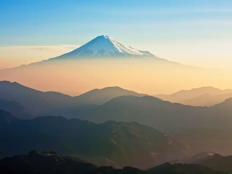 Der schneebedeckte Gipfel des Vulkans Fuji-san ragt über einer nebligen Berglandschaft in Japan bei Sonnenaufgang empor.