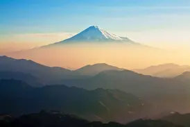 Der schneebedeckte Gipfel des Vulkans Fuji-san ragt über einer nebligen Berglandschaft in Japan bei Sonnenaufgang empor.