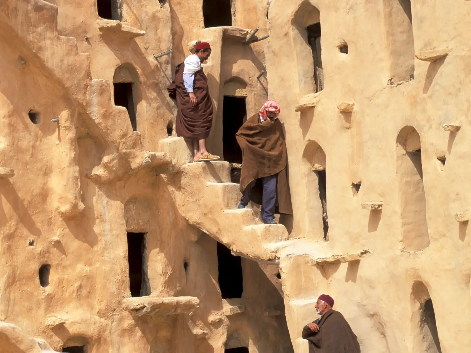 Einheimische Berber in traditioneller Kleidung auf einer Treppe an einem mehrstöckigen Lehmbau in der Region Matmata.