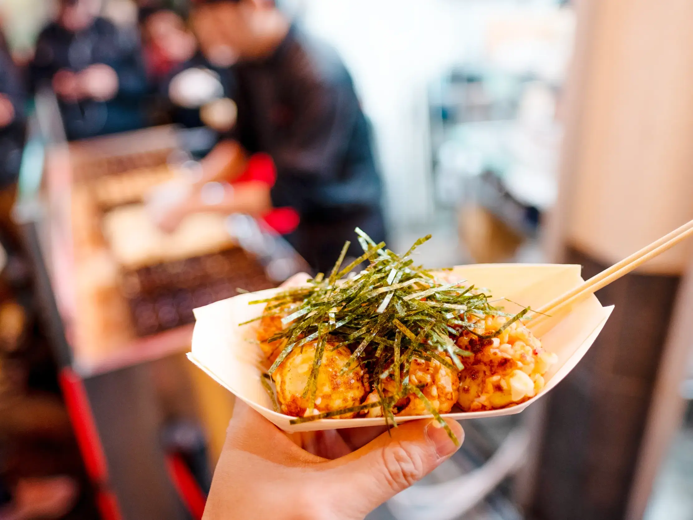 Eine Person hält eine Portion frischer Takoyaki mit Seetang in einer Papierschale auf einem Streetfood-Markt in Osaka, Japan.