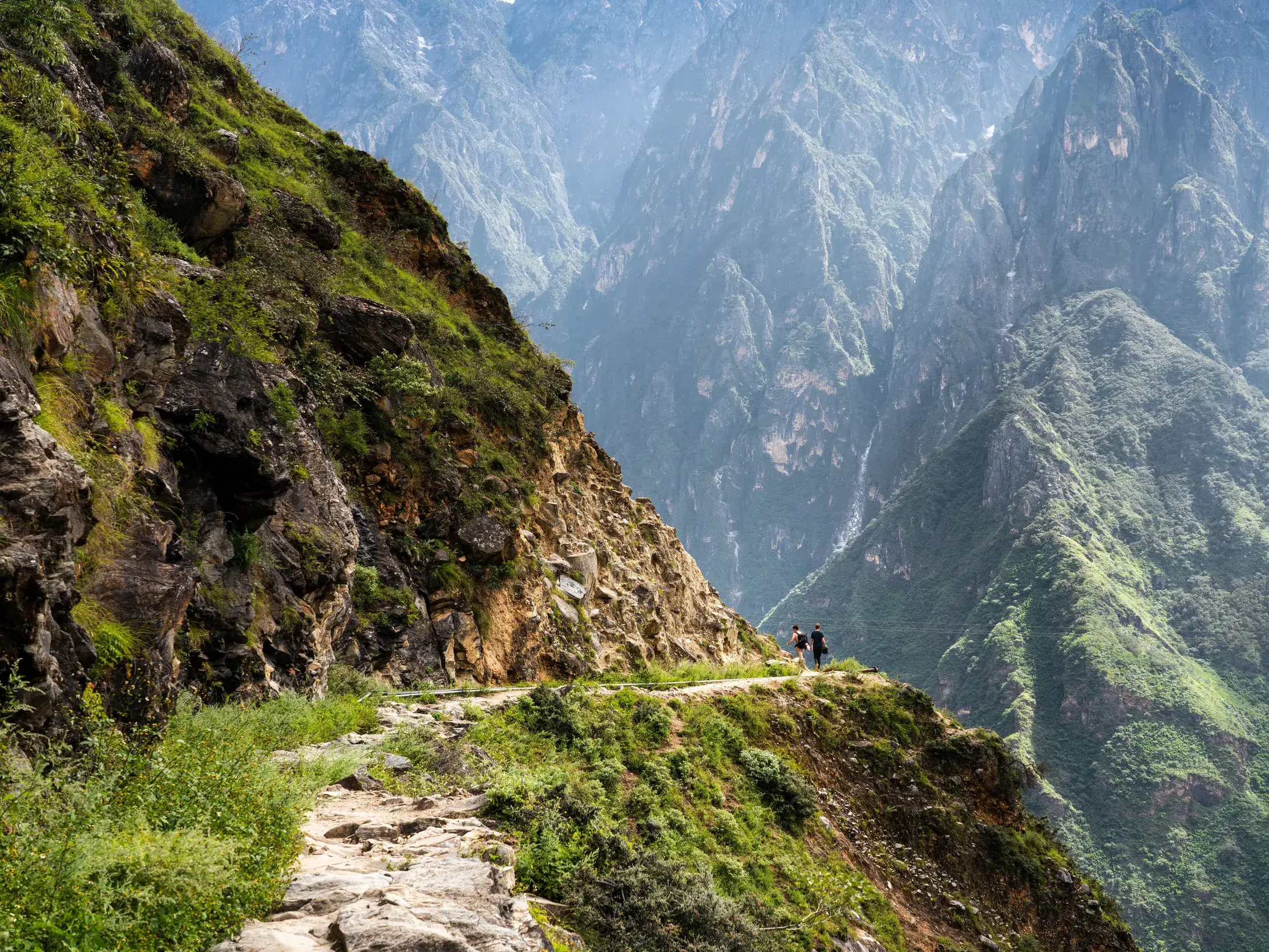 Zwei Wanderer auf einem schmalen Felsenpfad während einer Trekking-Tour durch die imposante Tigersprungschlucht in China.