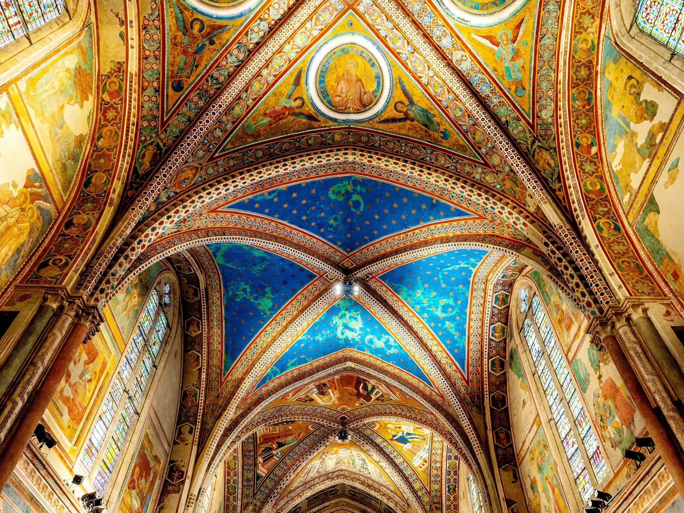 Das gotische Kreuzrippengewölbe der Basilika S. Francesco in Assisi, reich verziert mit farbenprächtigen Fresken und blauem Sternenhimmel.