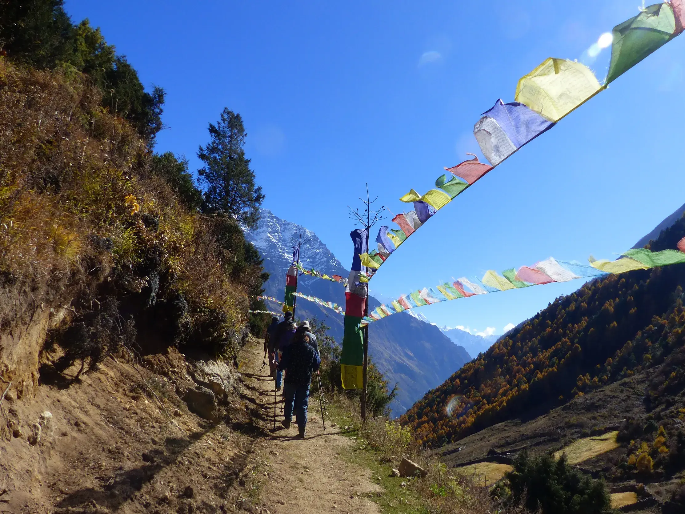 Eine Trekkinggruppe wandert auf einem Bergpfad in Nepal, eingerahmt von bunten Gebetsfahnen und mit Blick auf die Himalaya-Gipfel.