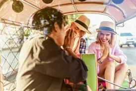 Lächelndes Paar mit Hüten bei einer Tuk-Tuk-Fahrt durch Bangkok, beide begrüßen den Fahrer mit der traditionellen Wai-Geste.