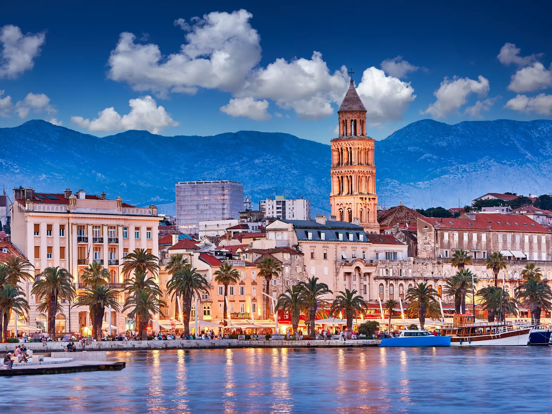 Die beleuchtete Uferpromenade Riva in Split bei Dämmerung, mit dem Diokletian-Palast vor der Bergkulisse Dalmatiens.