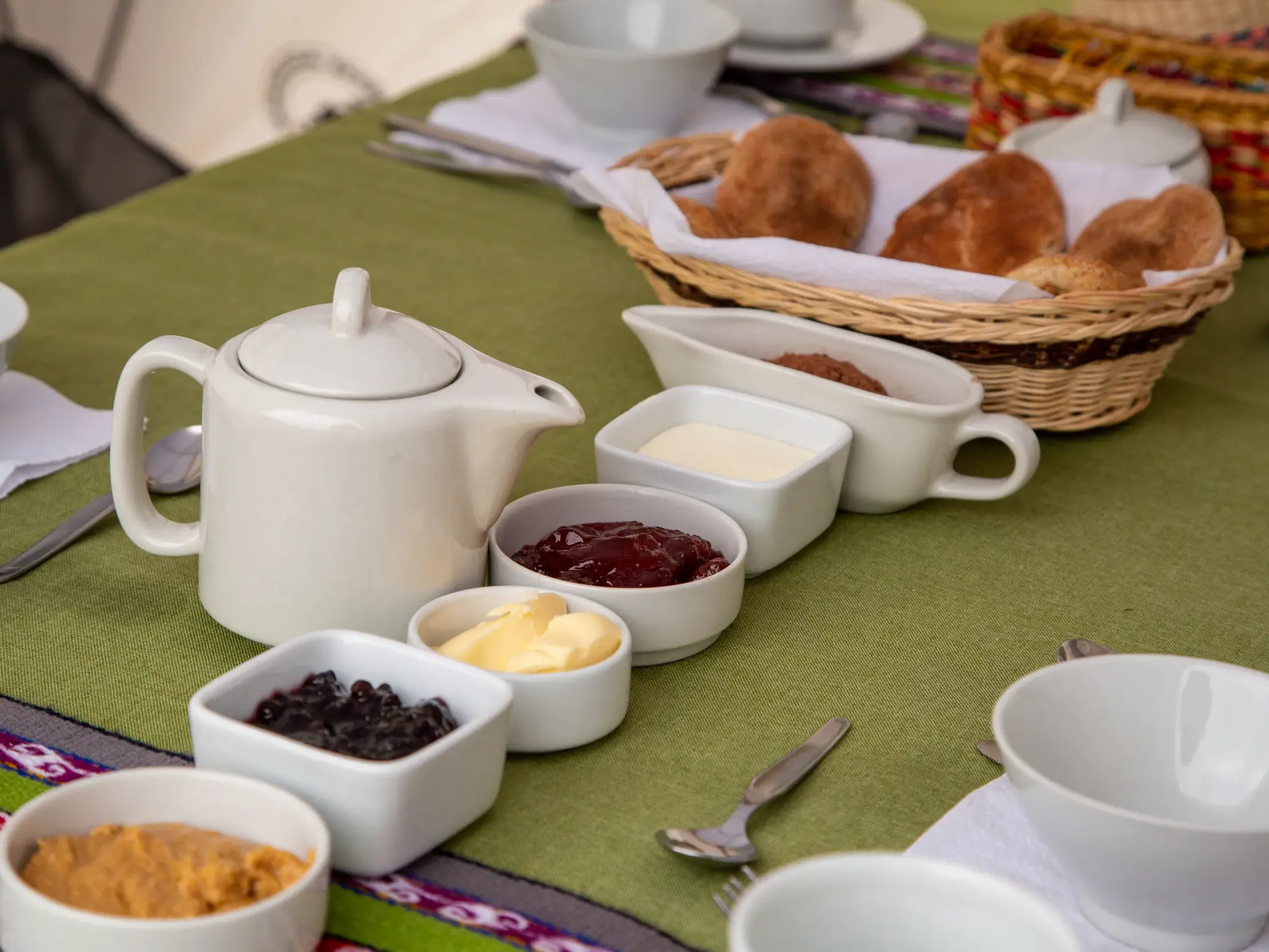 Liebevoll gedeckter Frühstückstisch mit Teekanne, Brotkorb und Aufstrichen auf einer grünen Decke mit peruanischem Muster.