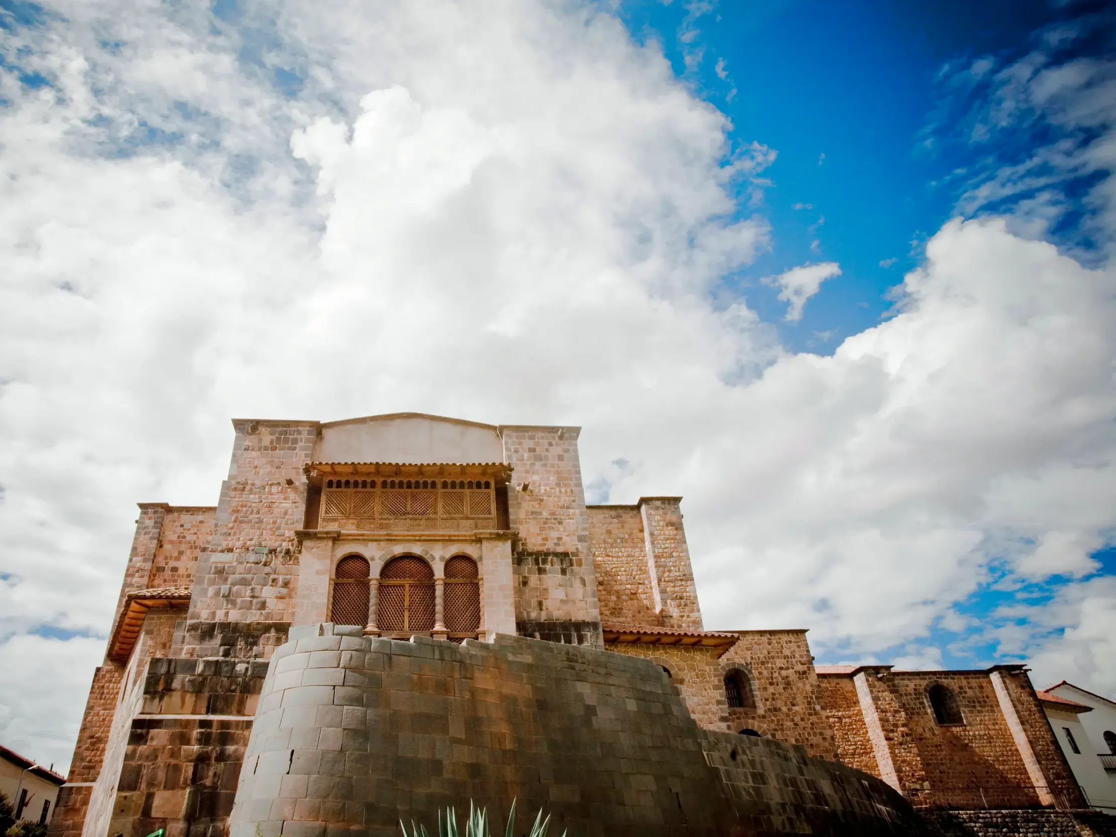 Froschperspektive eines historischen Gebäudes in Cusco mit massivem Inka-Fundament und Kolonialbau unter blau-weißem Wolkenhimmel.