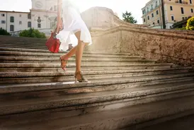 Eine elegant gekleidete Frau mit roter Tasche steigt bei Sonnenschein die historischen Stufen der Spanischen Treppe in Rom empor.