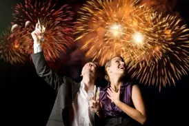 Zwei Reisende stoßen auf einer Singlereise in Lissabon zu Silvester fröhlich an, während ein Feuerwerk den Himmel erleuchtet.