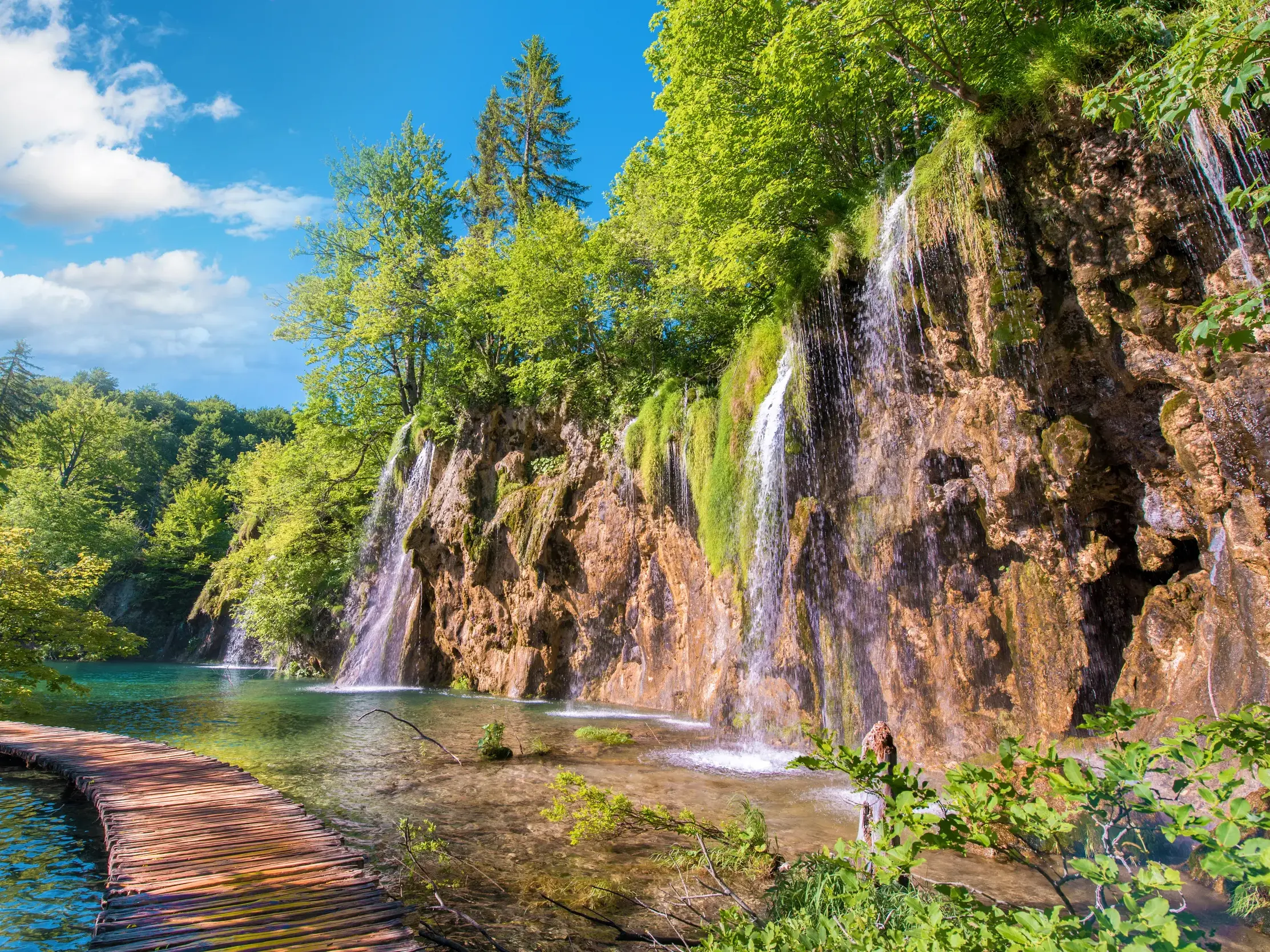 Ein hölzerner Wandersteg führt entlang türkisfarbener Seen und Wasserfälle im Nationalpark Plitvicer Seen in Kroatien.