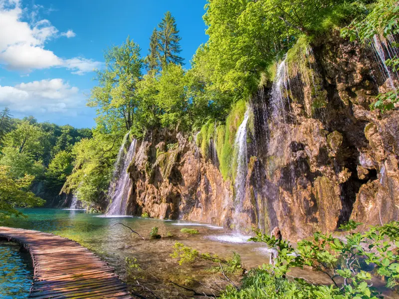 Ein hölzerner Wandersteg führt entlang türkisfarbener Seen und Wasserfälle im Nationalpark Plitvicer Seen in Kroatien.