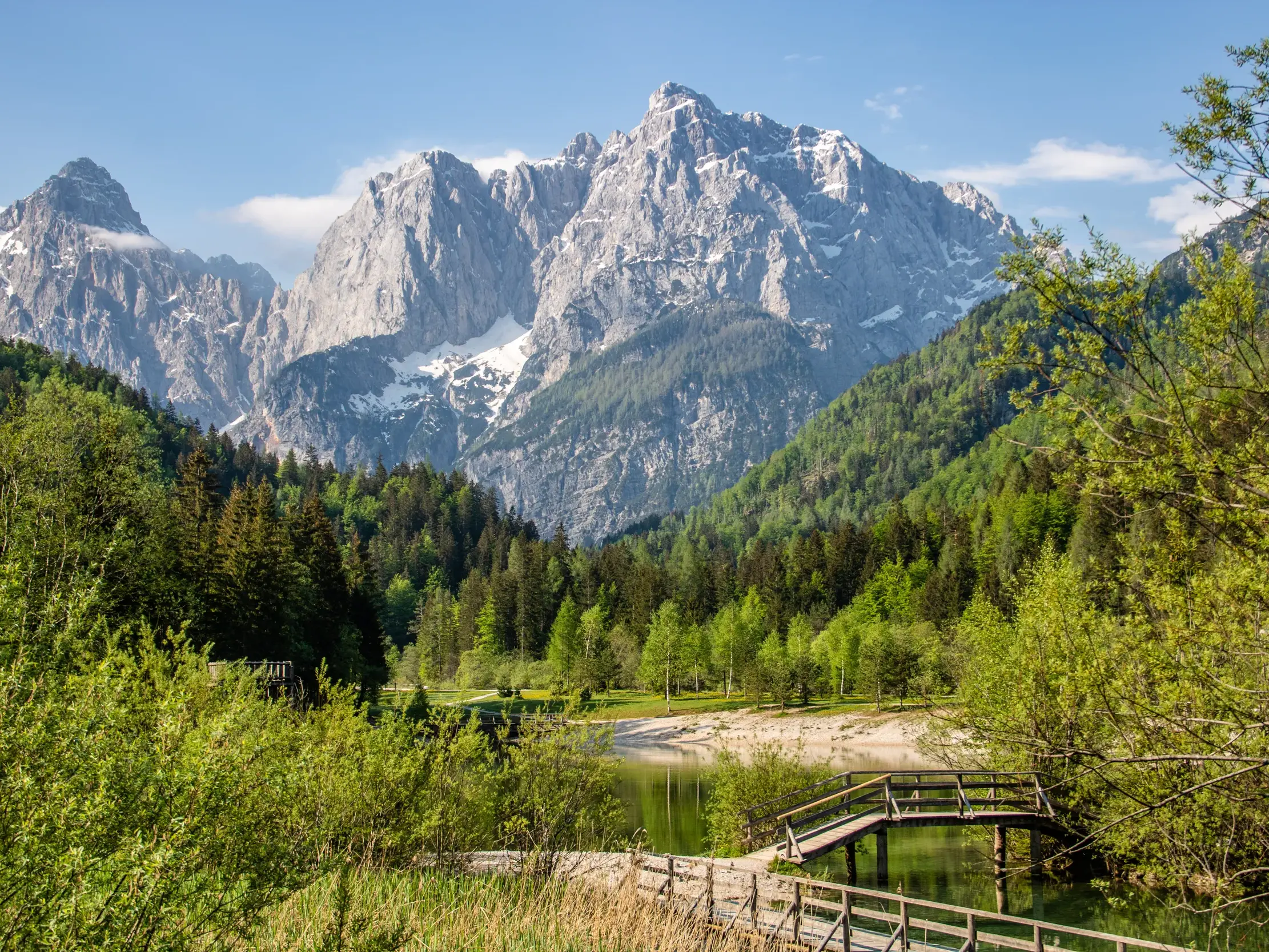Holzsteg am klaren Jasna See vor der Bergkulisse der schneebedeckten Julischen Alpen im Triglav-Nationalpark.