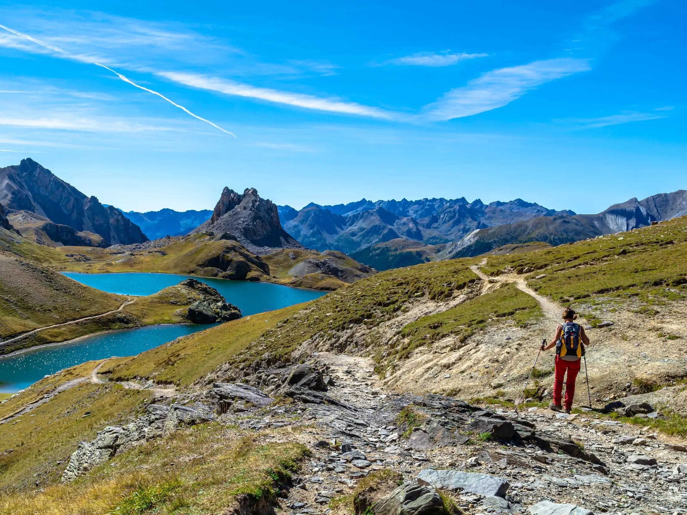Eine Wanderin genießt den Panoramablick auf zwei türkise Bergseen auf einem steinigen Wanderweg im Maira-Tal, Piemont.