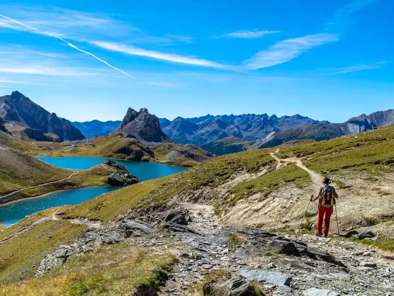 Eine Wanderin genießt den Panoramablick auf zwei türkise Bergseen auf einem steinigen Wanderweg im Maira-Tal, Piemont.