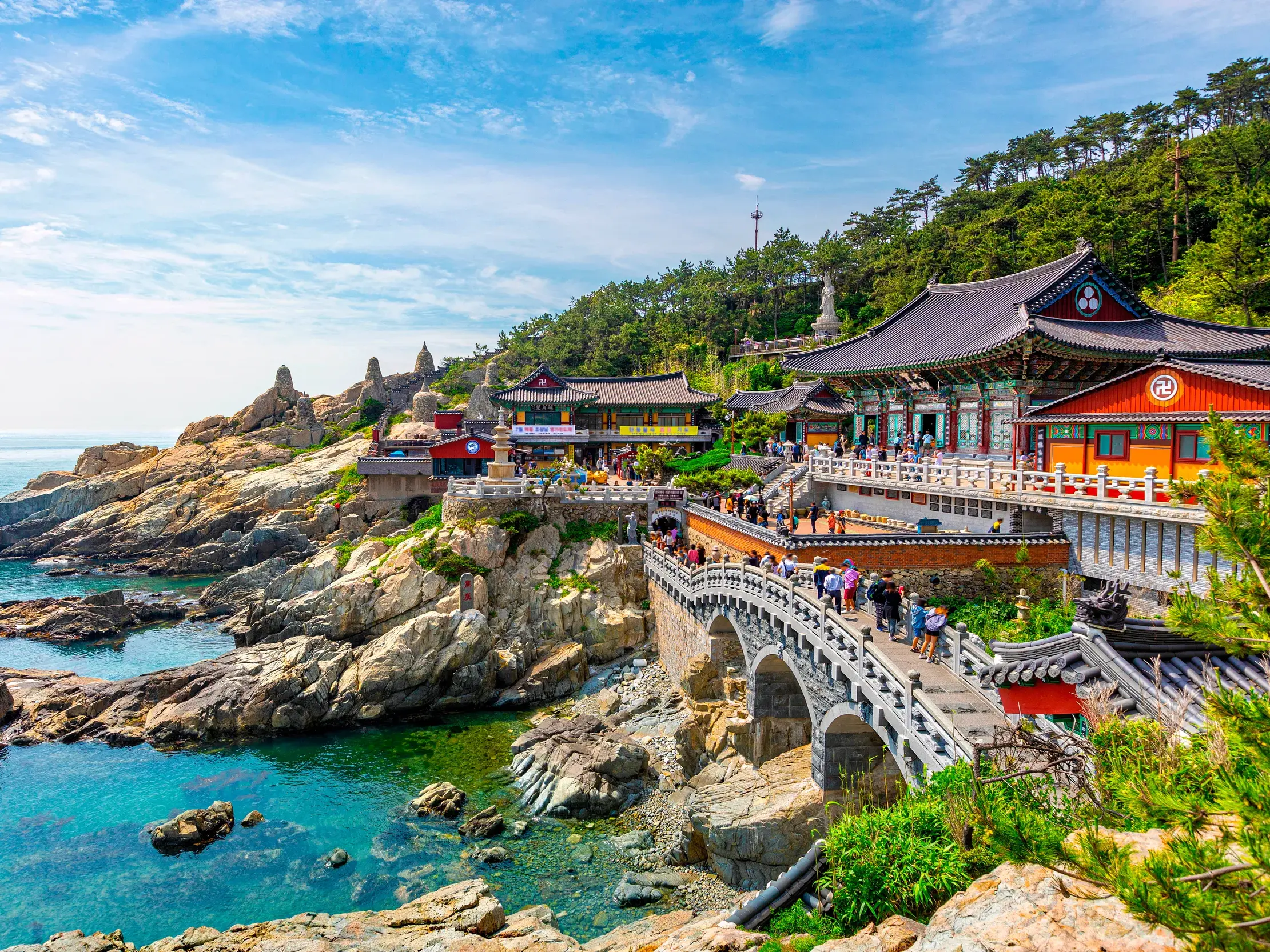 Bunte Tempelanlage Haedong Yonggungsa in Busan, malerisch auf Felsen am türkisen Meer erbaut, mit Besuchern auf einer Brücke.