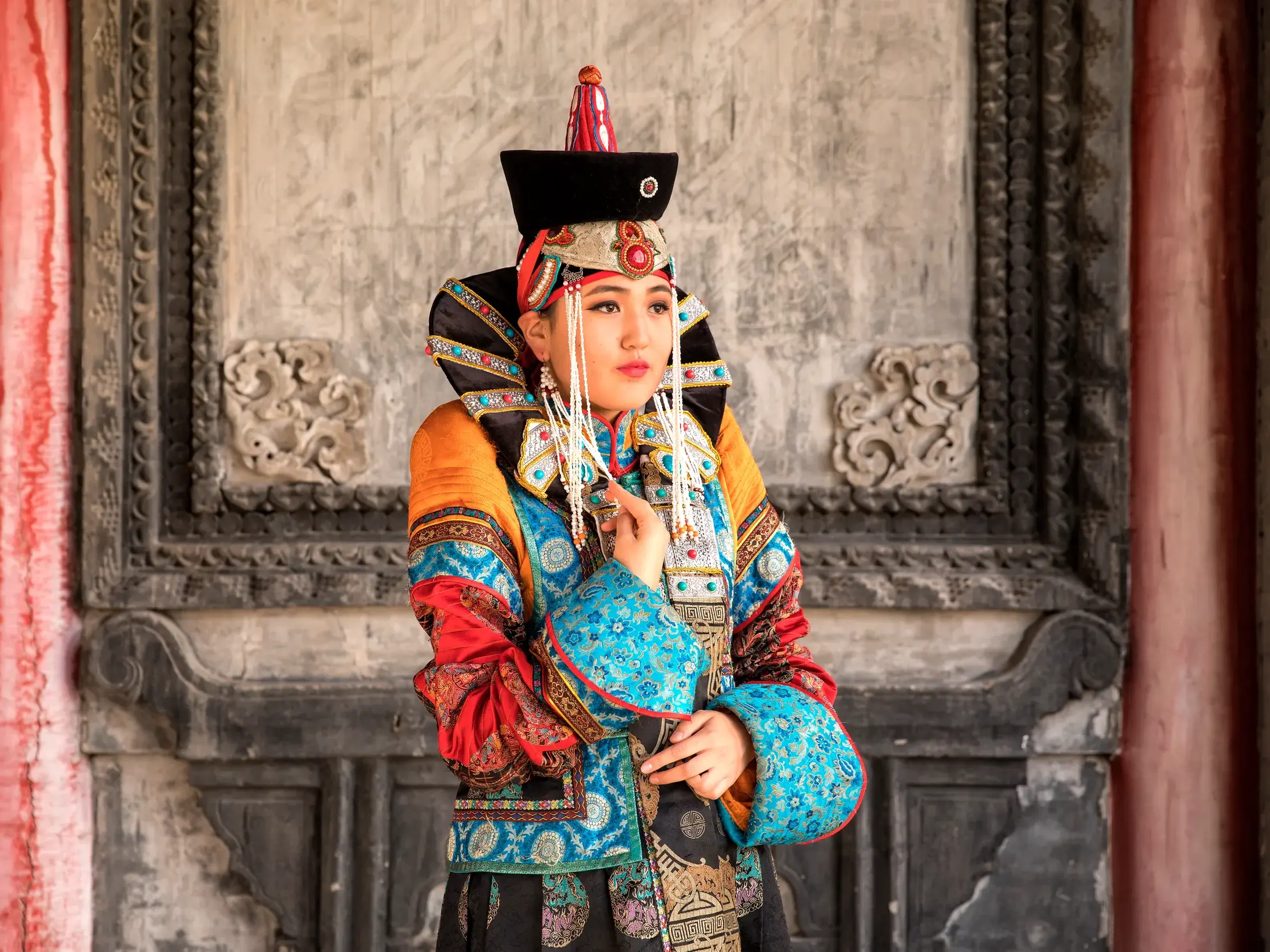 Junge Frau in einer farbenprächtigen, traditionellen mongolischen Tracht mit Kopfschmuck vor einer verzierten Tempelmauer in Ulan Bator.