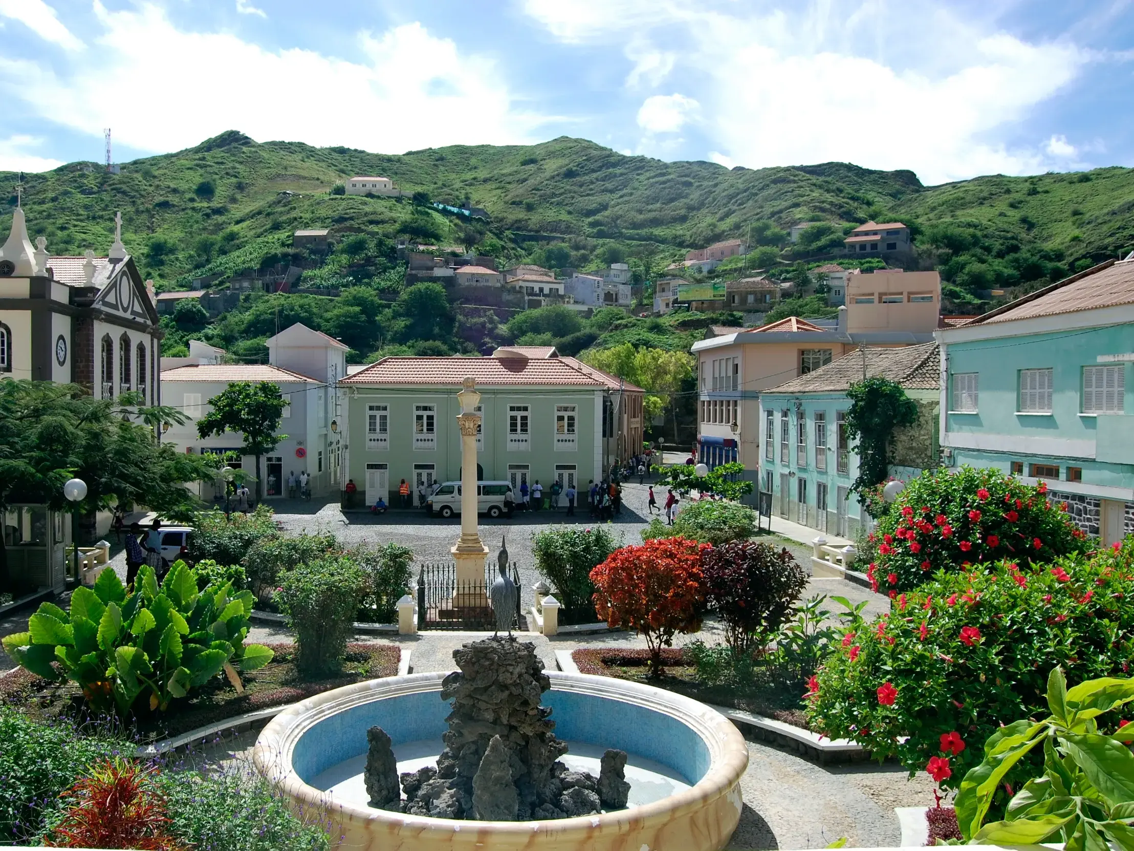 Blick auf den zentralen Platz von Ribeira Brava mit Brunnen, Kirche und bunten Häusern vor einem grünen Berghang auf São Nicolau.