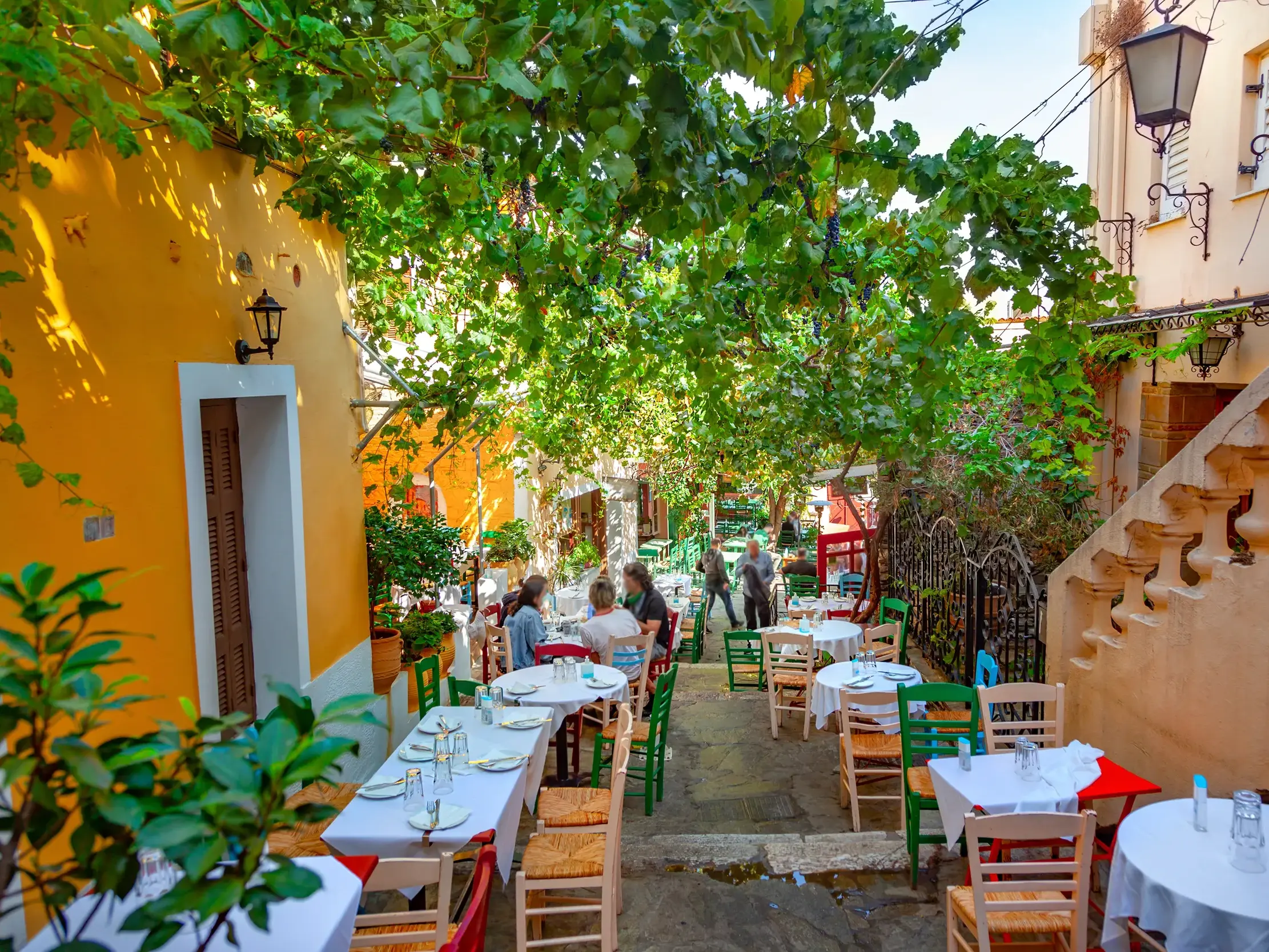Gedeckte Tische einer Taverne in einer sonnigen, von Weinreben überdachten Gasse in der Altstadt von Naxos in Griechenland.