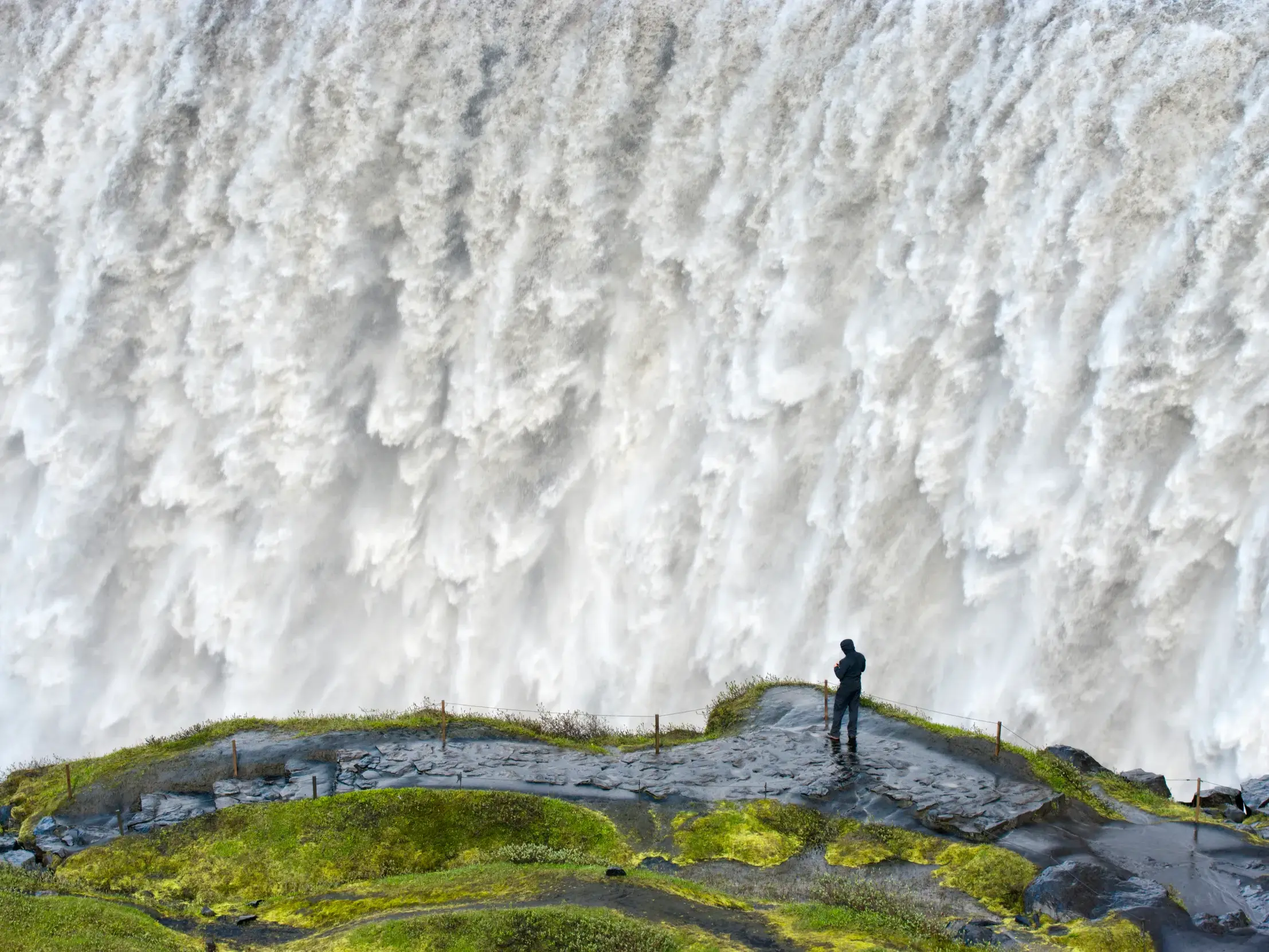 Eine Person steht auf einem moosbewachsenen Felsvorsprung und blickt auf die Urgewalt des Dettifoss Wasserfalls in Island.