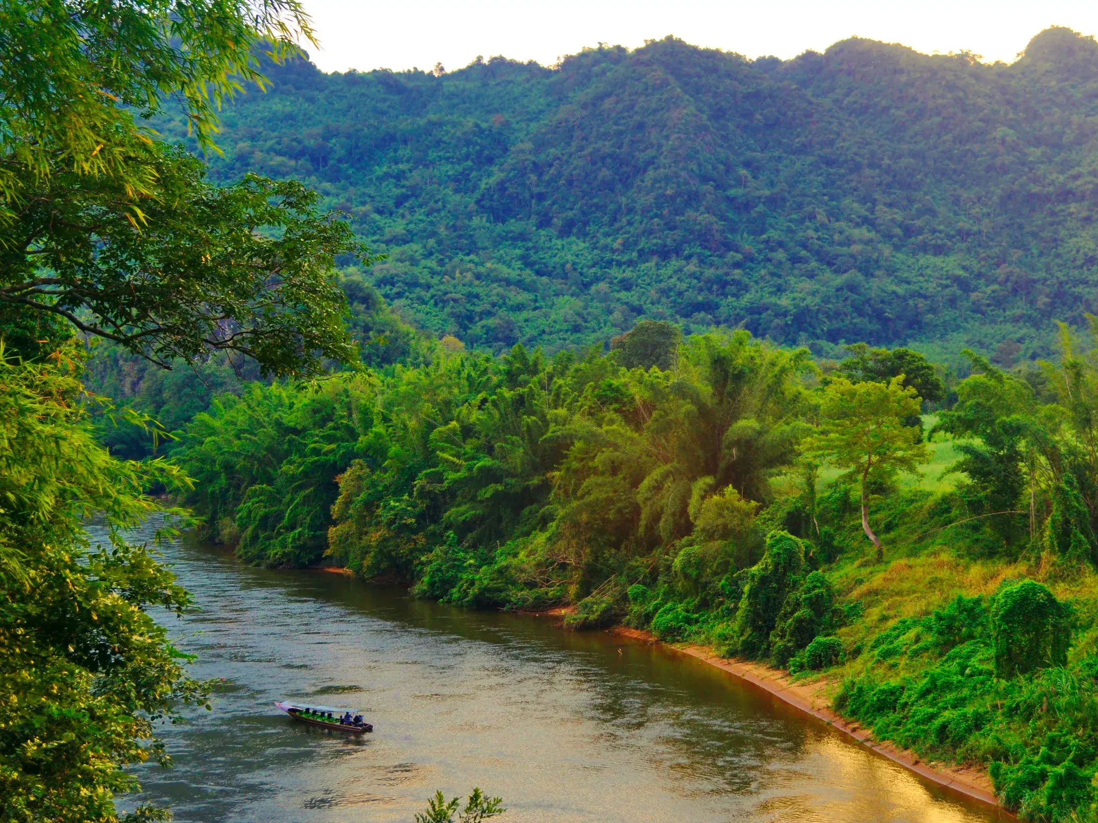 Ein Longtail-Boot mit Reisenden gleitet auf dem River Kwai durch die üppige, grüne Dschungellandschaft Thailands bei Kanchanaburi.