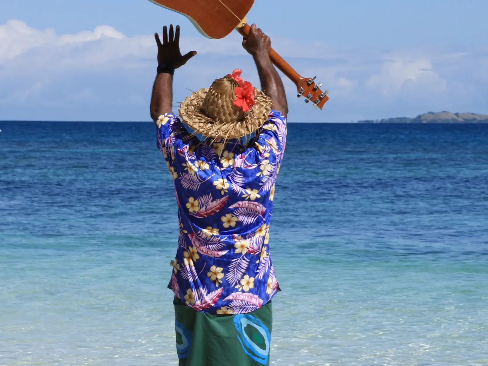 Einheimischer mit Ukulele und Blumenhemd begrüßt Reisende fröhlich am türkisblauen Meer auf einer der Inseln von Fiji.