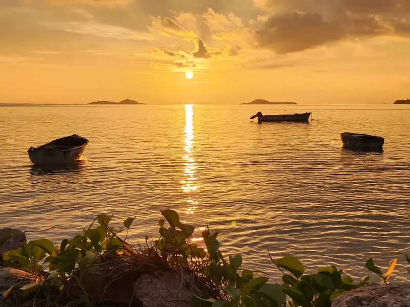 Drei kleine Boote ankern im Indischen Ozean vor der Küste der Seychellen bei einem stimmungsvollen, goldenen Sonnenuntergang.