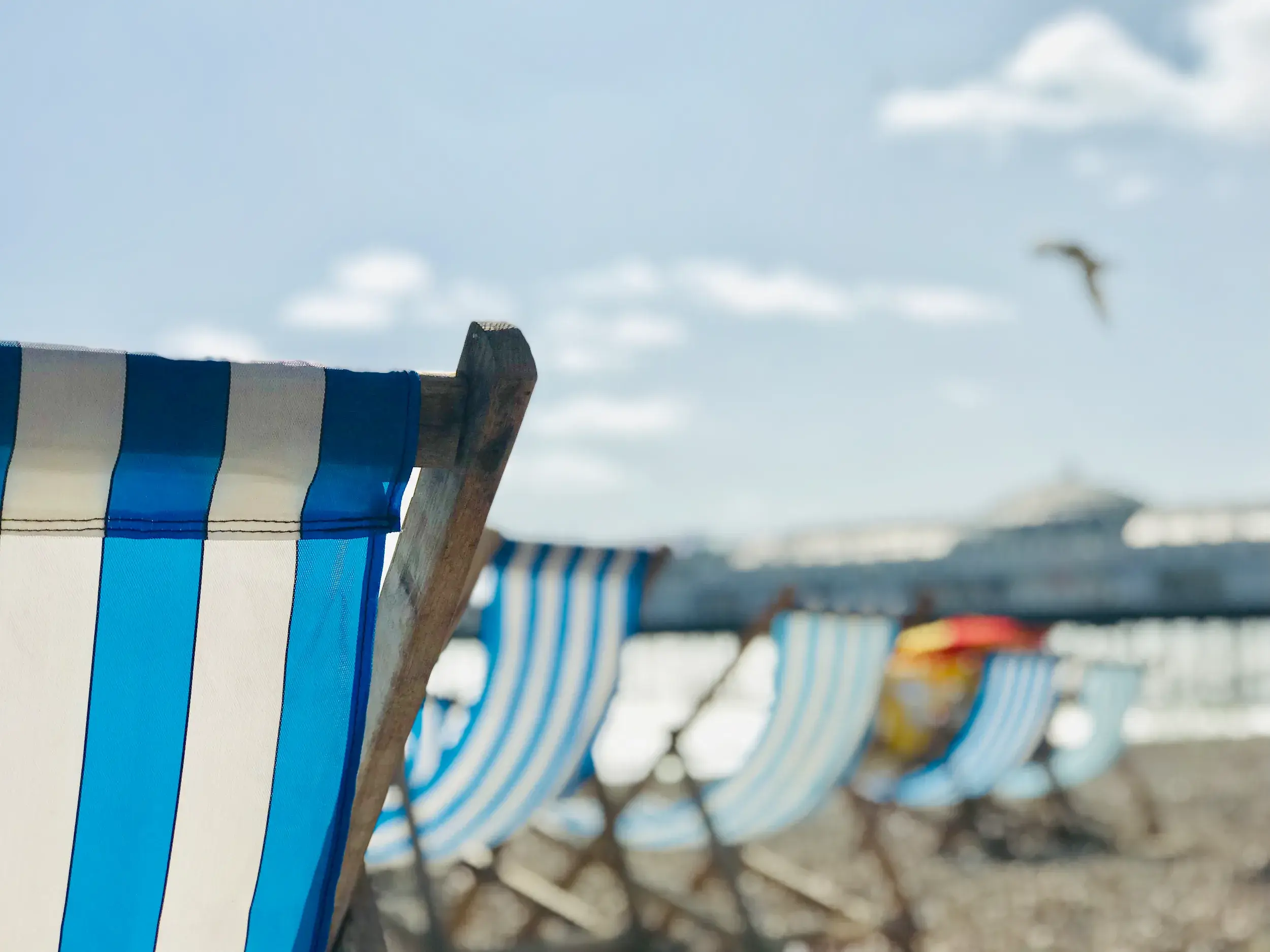 Nahaufnahme eines blau-weiß gestreiften Liegestuhls am Kieselstrand von Brighton vor der unscharfen Kulisse der Seebrücke.