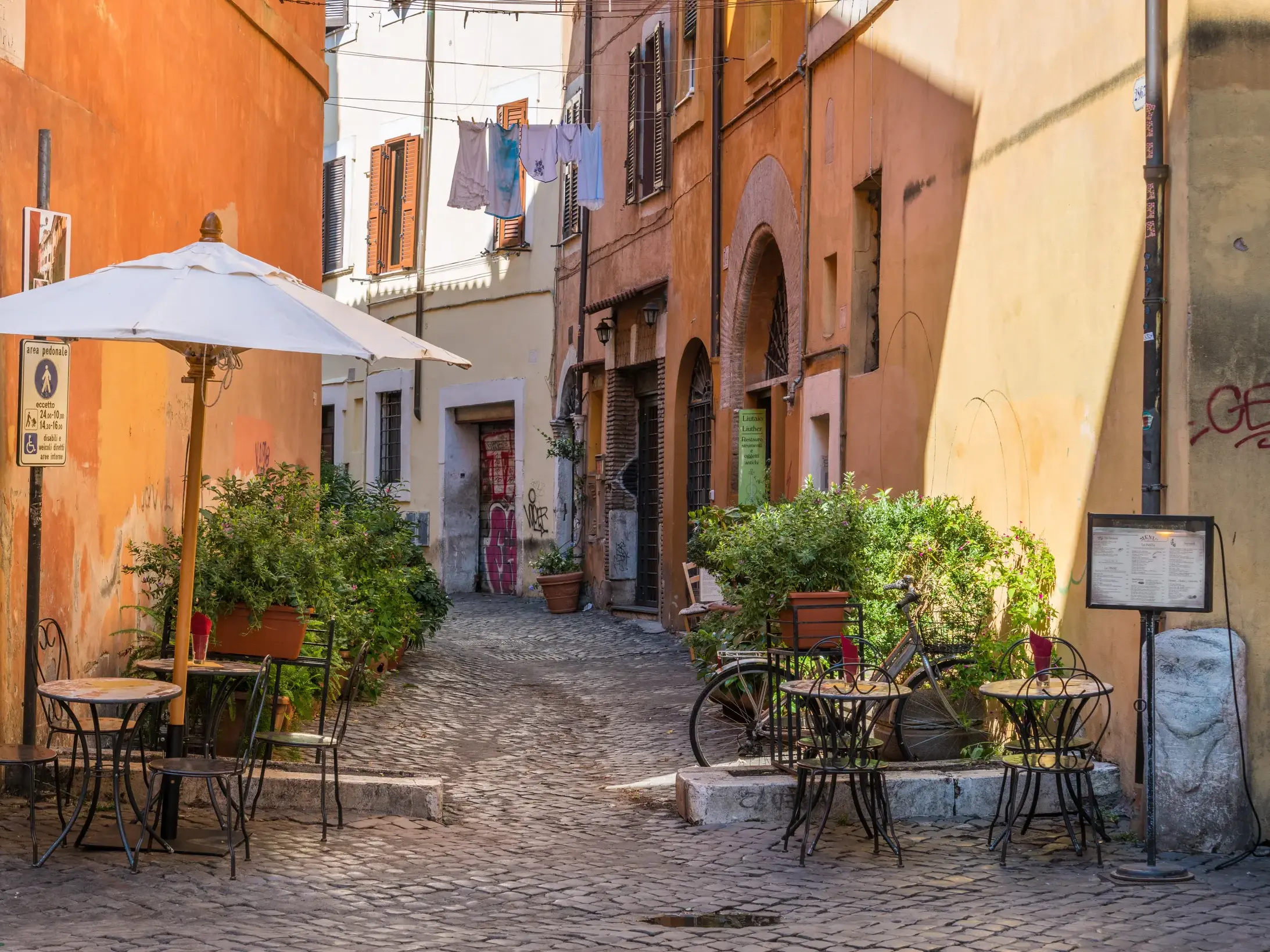 Sonnige Kopfsteinpflastergasse in Trastevere, Rom, mit Tischen und Stühlen eines Cafés, umgeben von ockerfarbenen Häusern.