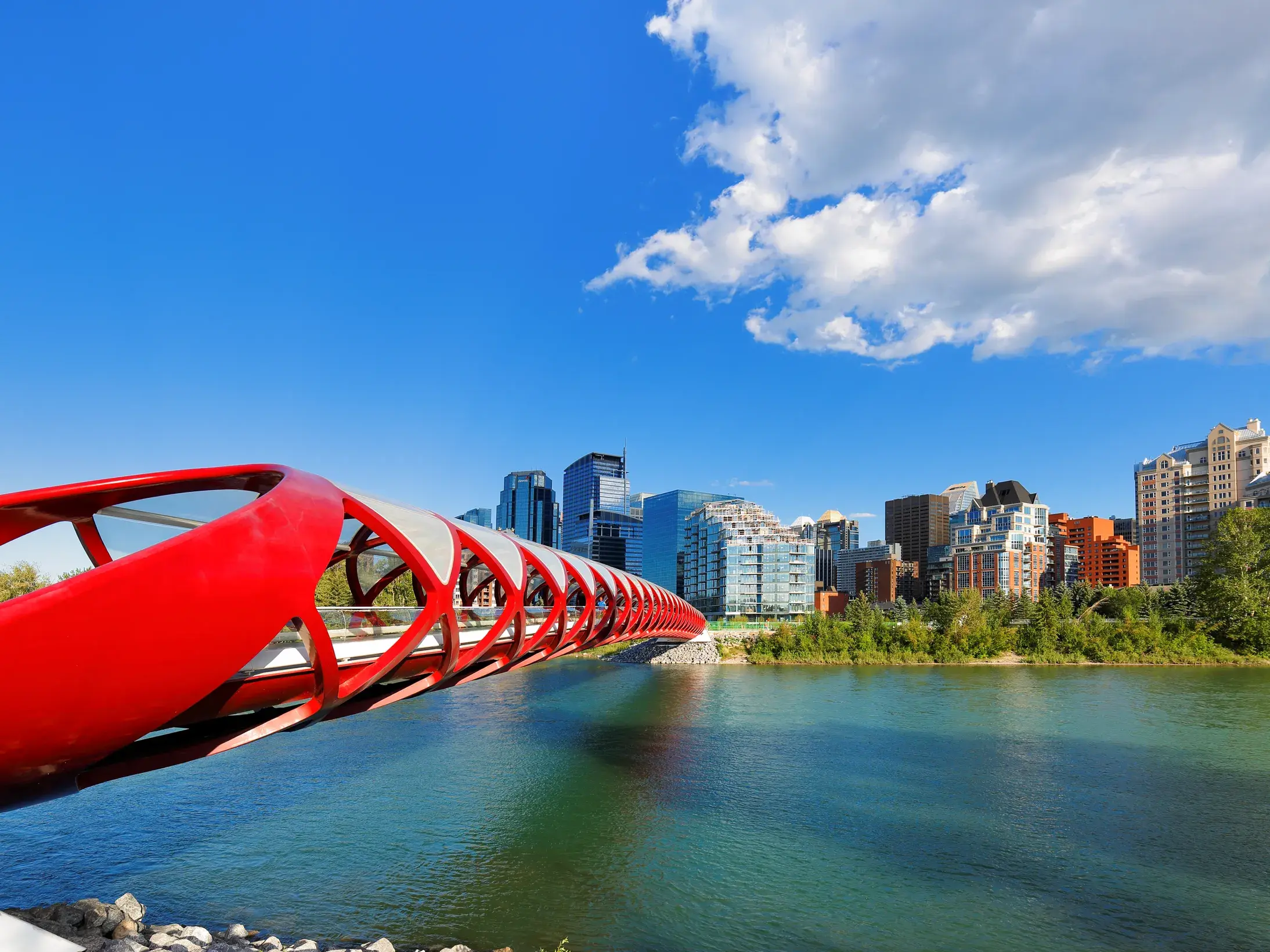 Die rote, röhrenförmige Peace Bridge für Fußgänger führt über den Bow River zur Skyline von Calgary bei Sonnenschein.