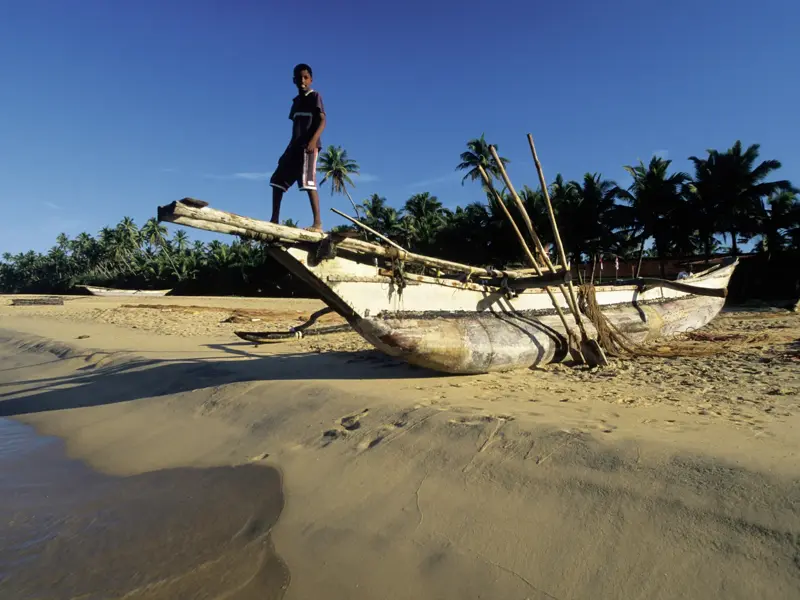 Einheimischer Junge steht auf einem traditionellen Auslegerkanu an einem Sandstrand an der Küste Sri Lankas bei Sonnenschein.