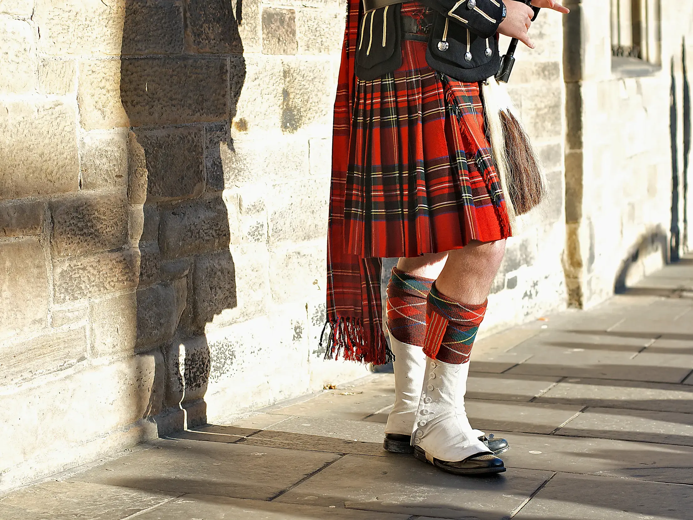 Nahaufnahme eines Mannes in traditioneller schottischer Tracht mit rotem Tartan-Kilt, der in den Highlands an einer Steinmauer steht.