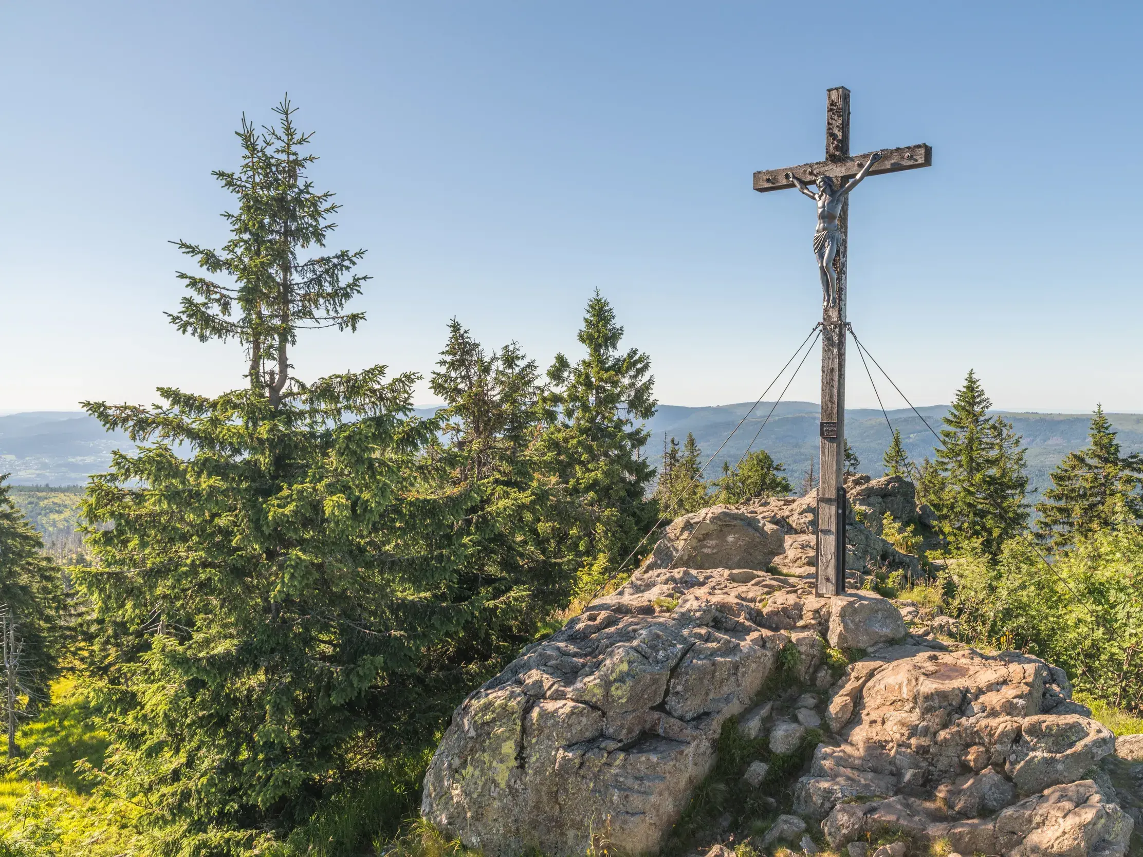 Majestätisches Gipfelkreuz auf einem felsigen Berggipfel, umgeben von Nadelbäumen, mit weitem Ausblick über eine grüne Berglandschaft.