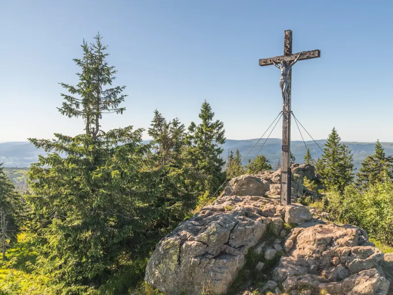 Majestätisches Gipfelkreuz auf einem felsigen Berggipfel, umgeben von Nadelbäumen, mit weitem Ausblick über eine grüne Berglandschaft.