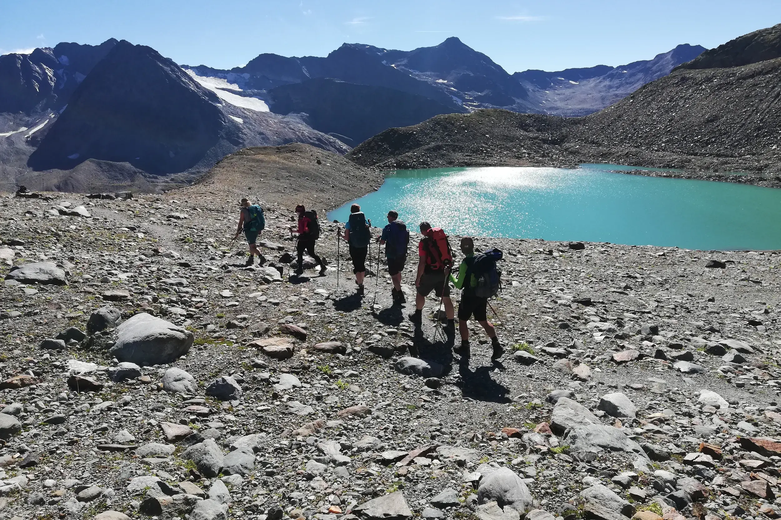 Eine kleine Wandergruppe wandert auf einem steinigen Weg an einem türkisen Gletschersee in den Stubaier Alpen vorbei.