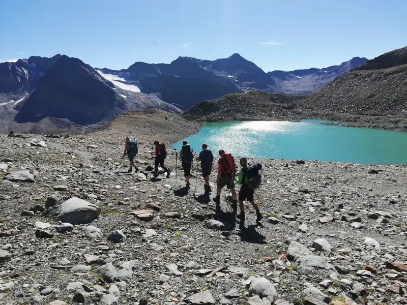 Eine kleine Wandergruppe wandert auf einem steinigen Weg an einem türkisen Gletschersee in den Stubaier Alpen vorbei.