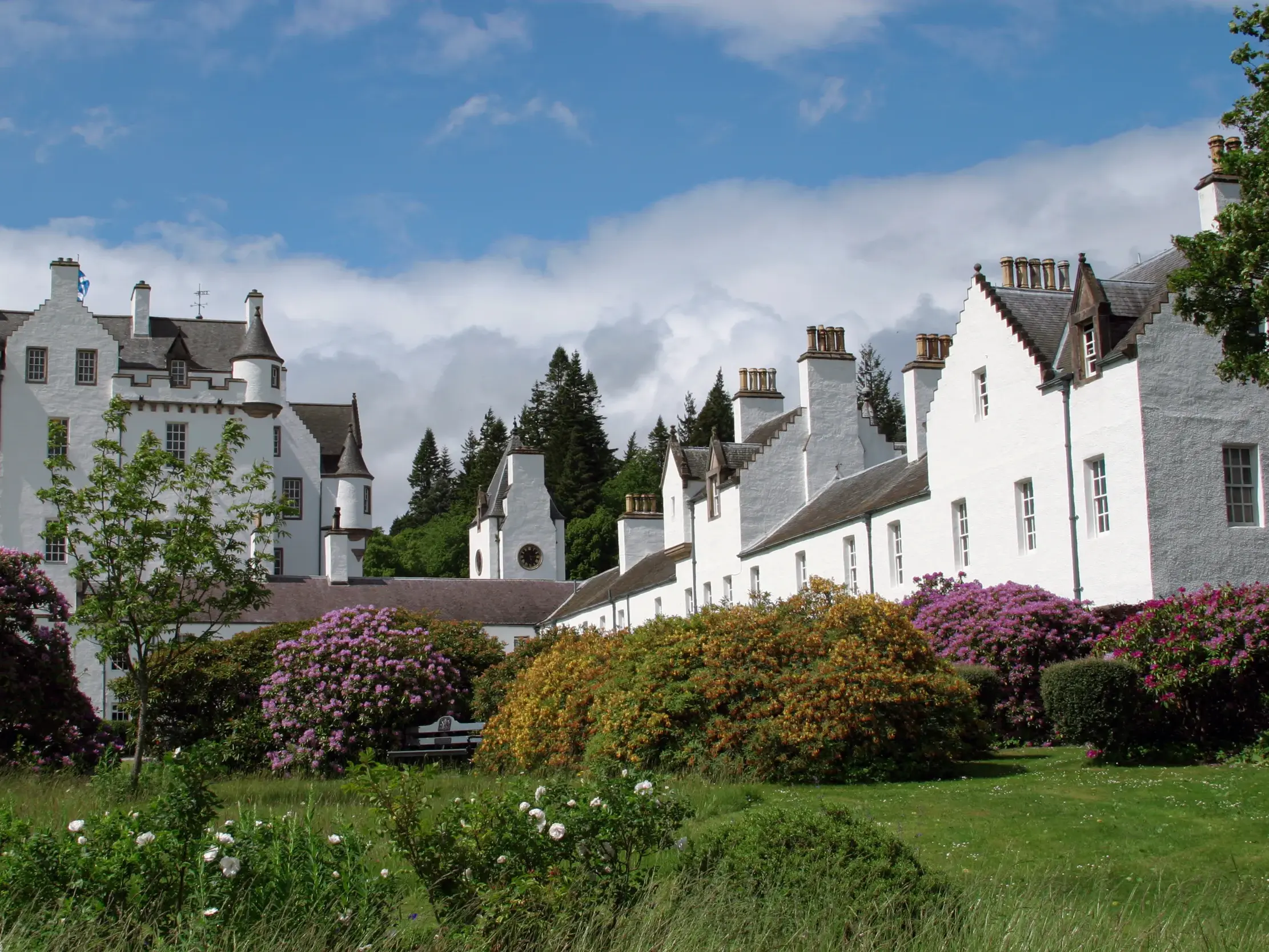 Das strahlend weiße Blair Castle in Schottland, umgeben von einem Park mit blühenden Rhododendren unter wolkigem Himmel.