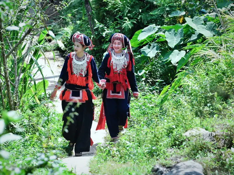 Zwei lächelnde Frauen des Bergvolks der Roten Dao in farbenfroher Tracht wandern auf einem Weg durch den Dschungel Vietnams.