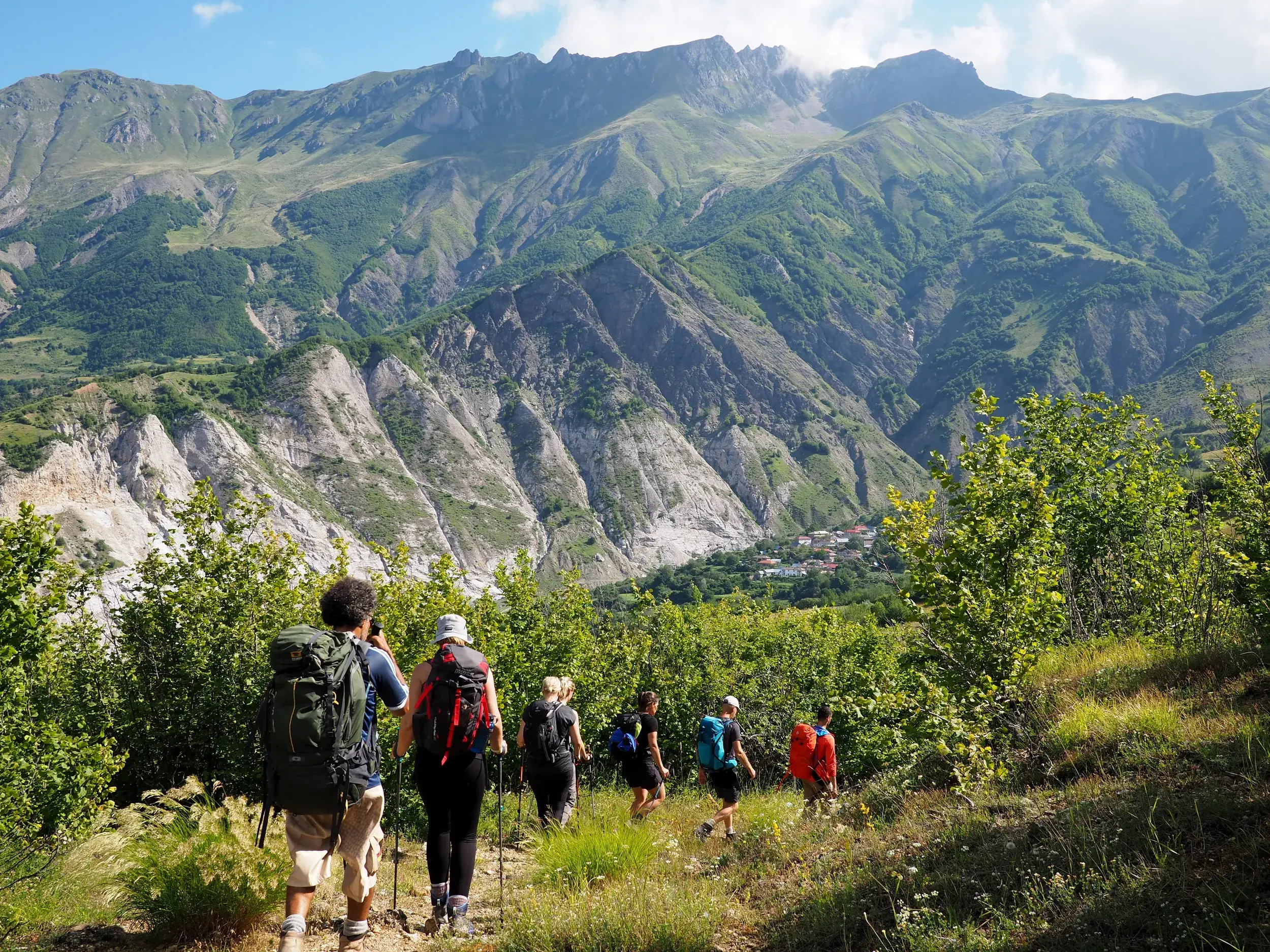 Hauser Reisegruppe beim Trekking auf einem sonnigen Pfad im Korab-Gebirge in Albanien mit Blick auf ein Bergdorf.